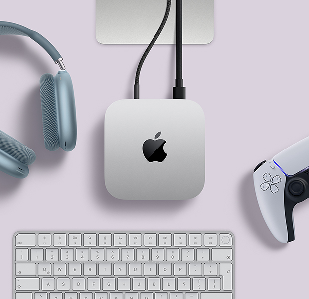 Vista desde arriba de un Mac mini con dos cables conectados en los puertos traseros. Junto al Mac mini hay unos AirPods Max azules, un Magic Keyboard color plata y un control inalámbrico para jugar