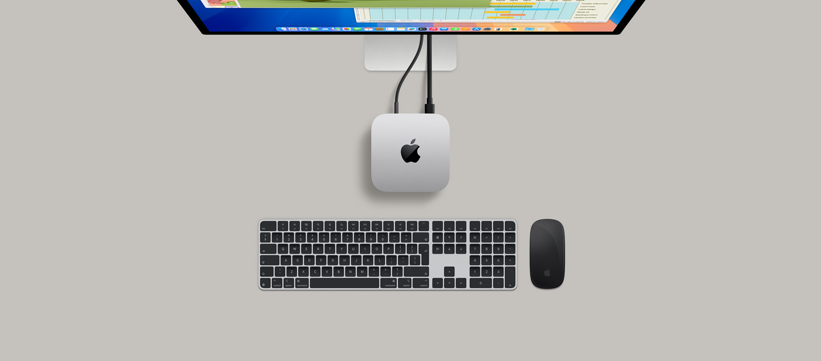 Top view of a silver Mac mini plugged into a display showing a game in active play, and next to a wireless keyboard, a gaming controller, and blue AirPods Max