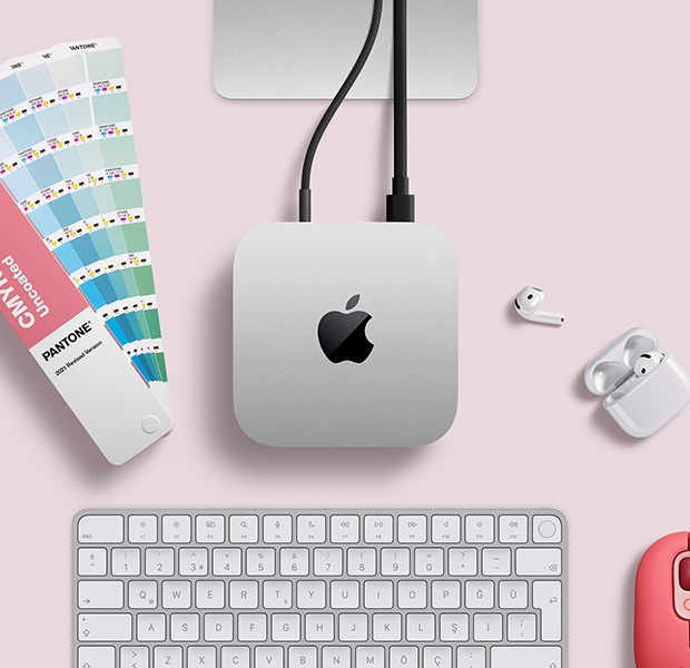 AirPods, pembe bir kablosuz klavye, pembe bir kablosuz mouse ve açık duran bir renk kartelası ile birlikte masa düzeninde yer alan gümüş rengi Mac mini'nin üstten görünümü