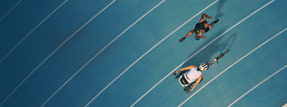 Duas atletas se enfrentam na pista de corrida. Uma está correndo com as próprias pernas e a outra está em uma cadeira de rodas adaptada para corridas.