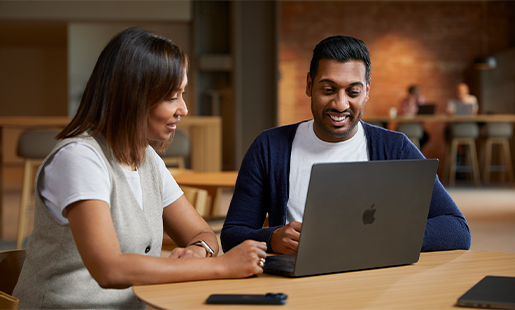 Amar seduto a un tavolo di uno spazio comune mentre usa un MacBook e collabora con una collega.