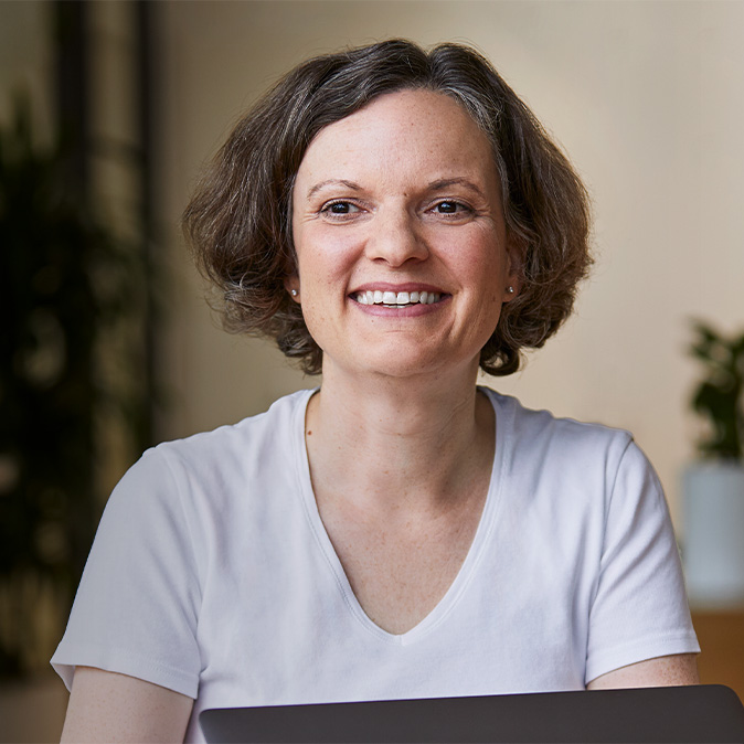 Ruth smiling, sitting indoors with plants in the background.