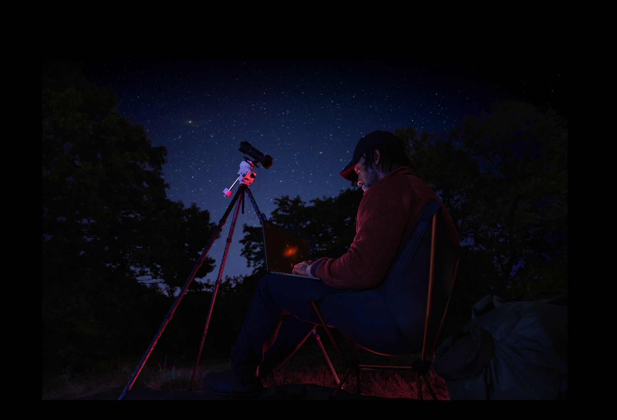 A person outside at night using a MacBook Pro on their lap, connected to a device on a tripod pointed towards the starry sky